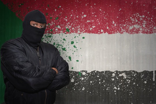 dangerous man in a mask standing near a wall with painted national flag of sudan