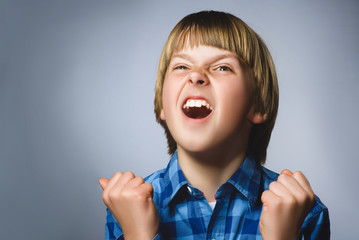Portrait of angry boy in blue shirt with hands up yelling isolated on gray studio background. Negative human emotion, facial expression. Closeup