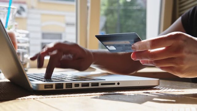 Woman makes purchases with a credit card online with a laptop sitting in a cafe near the window. Hands, credit card and fingers on the keyboard close-up