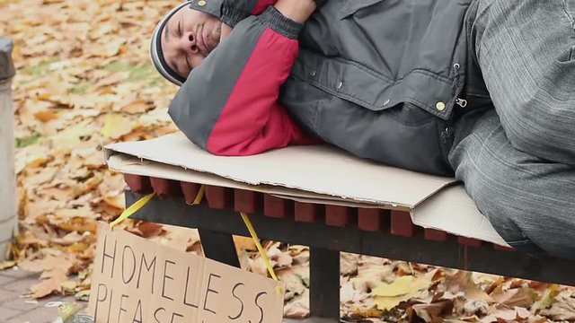 Man dressed in old clothes sleeping on bench in park with homeless help sign