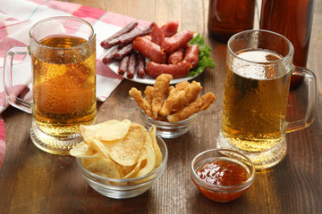 light beer in glass and in a glass bottle with a variety of sausages in a dish near the bowl of chips and ketchup on wooden brown background