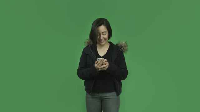 caucasian girl with winter jacket isolated on green screen smiling with cell phone