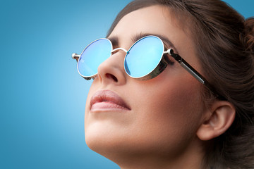 Close-up face portrait of young beautiful woman with perfect skin in round sunglasses looking up on blue background. Beauty face make-up. 