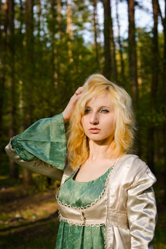  Fashionable shooting of a young short haired blond woman posing in the forest park wearing fancy empire style dress holding flowers. Concept of fantasy and magic.