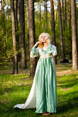 Obraz na płótnie Canvas Fashionable shooting of a young short haired blond woman posing in the forest park wearing fancy empire style dress holding flowers. Concept of fantasy and magic.