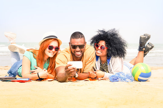 Multiracial friends having fun taking selfie lying on the beach - Happy multiethnic students watching self photo on vacation - Concept of teens joyful moment and technology addiction    