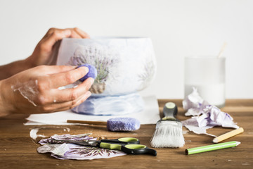 Decoupage hobbyist hands decorating a vase with lavender pattern - some artistic supplies on a table. 
