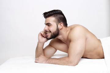 Junger Mann mit Bart liegt nackt auf Massage Liege und lächelt