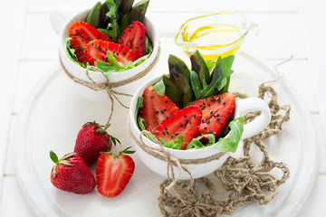 Fresh salad with strawberry, arugula, avocado and black sesame in white cups on white wooden background