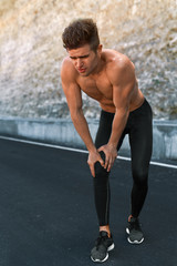 Obraz premium Sport Injury. Athletic Man With Fit Muscular Body Holding Painful Knee And Feeling Ache, Suffering From Pain. Handsome Fitness Runner Injured His Leg During Running Workout Training Outdoors.
