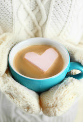 Female hands in warm mittens holding cup of hot cappuccino with heart marshmallow, close up