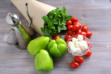 Wet fresh basil (herbs) in the cone of paper, cherry tomatoes, green sweet pepper, mozzarella cheese and oil jug on a wooden table