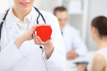 Female doctor with stethoscope holding heart.  Doctor and patient sitting in the background