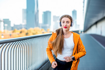 Young sport woman in yellow sweater with earphones running on the modern bridge with skyscrapers on...
