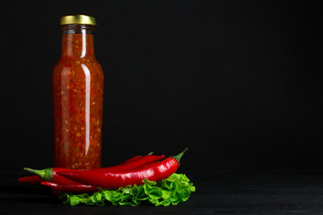 red sauce in a glass bottle near the red hot pepper and greens leaves on black wooden background