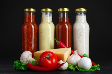 a variety of sauces in glass bottles with vegetables and spices on a black wooden background