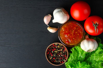 red sauce in a glass bowl near the red hot peppers and the leaves are green with vegetables on a black wooden background