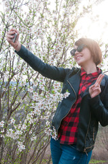 Lifestyle portrait of fashionable woman wearing stylish jacket, checkered shirt, jeans, glasses and taking selfie 