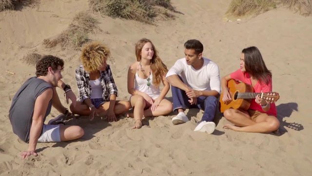 Group of young multiracial friends sitting on a sandy beach playing guitar together and singing and chatting in the evening sun