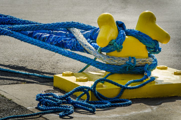 Heavy blue ropes of an ocean-going ship wrap around a yellow mooring bollard on a city pier in the harbor. - 111203576