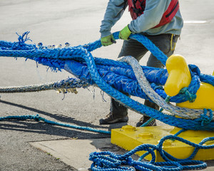 Dock worker pulls on heavy blue ropes of an ocean-going passenger ship are wrapped around a yellow mooring bollard on a city pier in the harbor. - 111203716