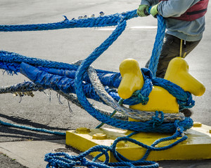 Dock worker pulls on heavy blue ropes of an ocean-going passenger ship are wrapped around a yellow mooring bollard on a city pier in the harbor. - 111203788