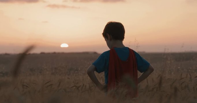 Boy with a superman cape stands in a golden wheat field, raising his hands in victory