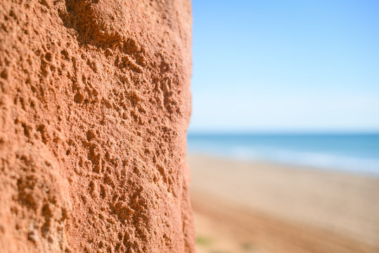 Closeup on red rock wall with defocused beach and sea. Sunny outdoors background
