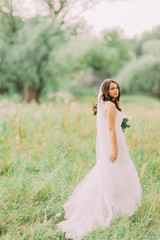 Fototapeta na wymiar Portrait of beautiful young bride in elegant dress with long veil and white flowers bouquet in the park