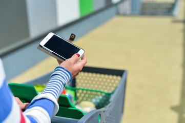 Closeup on person holding mobile smart phone in hand during shopping. Cart on store background
