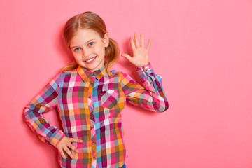 Hello! Pretty little girl standing with raised hand, smiling and welcomes you on pink background. Copy space.