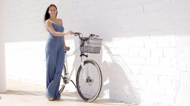 Elegant young woman in a denim slack suit standing holding a bicycle against a white wall in the summer sunshine