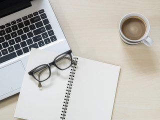 Eyeglasses on notebook, laptop computer and a cup of hot coffee on wooden table