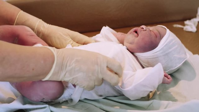 Nurse put on white disposable diaper on newborn crying baby in hat after birth. Maternity hospital.