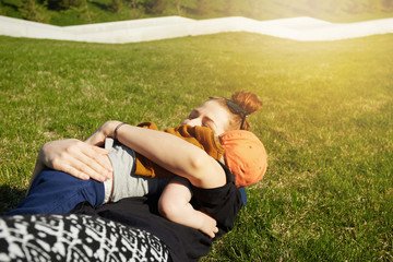 Portrait of happy young mother spending great time with her adorable two-year old son, hugging him in tenderness while lying on the grass in the public garden during walk in the morning. Flare sun