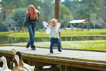 Kid girl and mother playing with ducks in lake