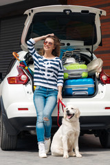 Beautiful young woman in sunglasses, wearing a t-shirt with white and dark blue stripes,blue jeans with holes in the knees,standing with his white dog near the white car, loaded with stuff