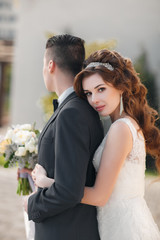 Young loving couple,the groom in a dark suit, bride in beautiful wedding dress,a brunette with long curly hair and a diadem,standing with a bouquet of flowers embracing on the street