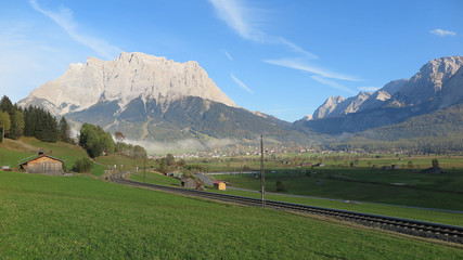 Fototapeta na wymiar Idyllic scenery of Tyrolean countryside with railway tracks crossing green fields in the foreground and rugged Zugspitze Mountain in the background on a beautiful sunny day in Lermoos, Tirol, Austria