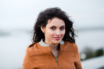 Close-up portrait of young happy beautiful attractive woman wearing leather coat smiling and standing over light outdoors background. Wind blow her hair.