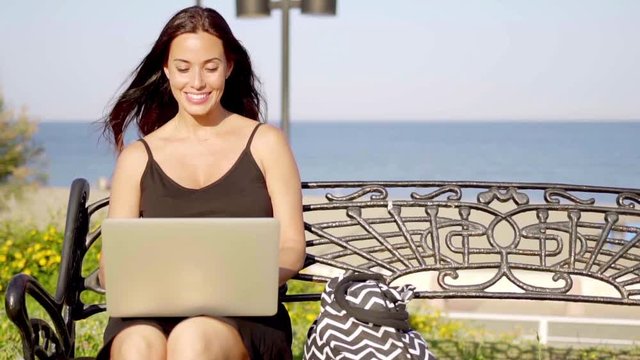 Attractive stylish young woman sitting on a wrought iron bench at the seaside typing on her laptop with a happy smile