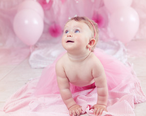 Obraz na płótnie Canvas Portrait of cute adorable Caucasian baby girl with blue eyes in pink tutu skirt celebrating her first birthday with gourmet cake and balloons looking away, cake smash first year concept