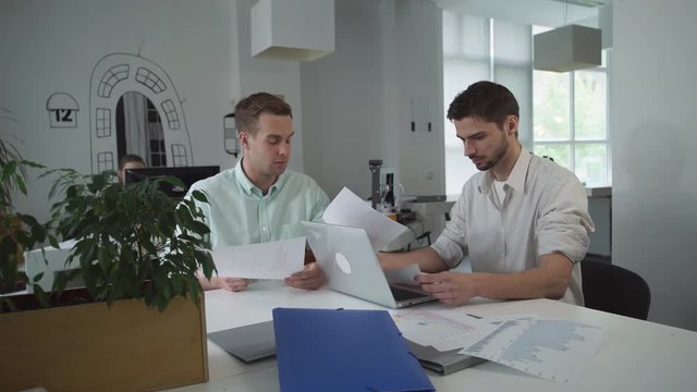 Casual young professional man holding paper report with diagram. Colleagues chatting together and discussing their work. 