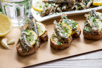 Snack from sandwiches with sardines. In the foreground sandwiches with fresh sardines, in the background a plate of sardines, a glass of water, parsley, garlic, lemon, olive oil. Horizontal. Daylight.