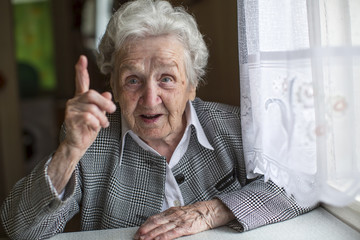 An elderly woman sitting at a table and specifies the finger. - obrazy, fototapety, plakaty