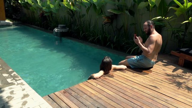 Couple relaxing by swimming pool, man taking photo of his girlfriend with cellphone

