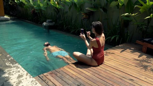 Couple relaxing by swimming pool, woman taking photo of her boyfriend with cellphone
