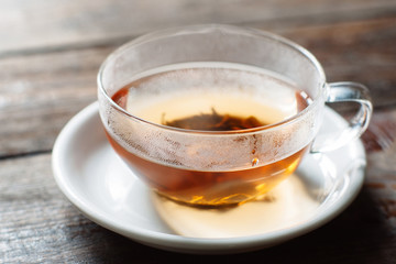 Cup of hot black tea close-up. Glass cup on saucer front view. Cup with tea leaves on dark wooden background. 