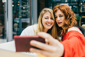 Half length of two young handsome caucasian blonde and redhead hair women outdoor in the city, holding smartphone, taking selfie, smiling - social network, communication, vanity concept