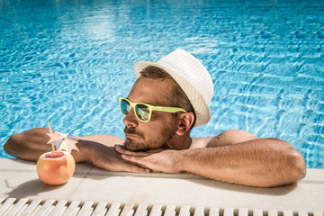 Portrait of a handsome man resting at the edge of a swimming pool. Model is wearing hat and...